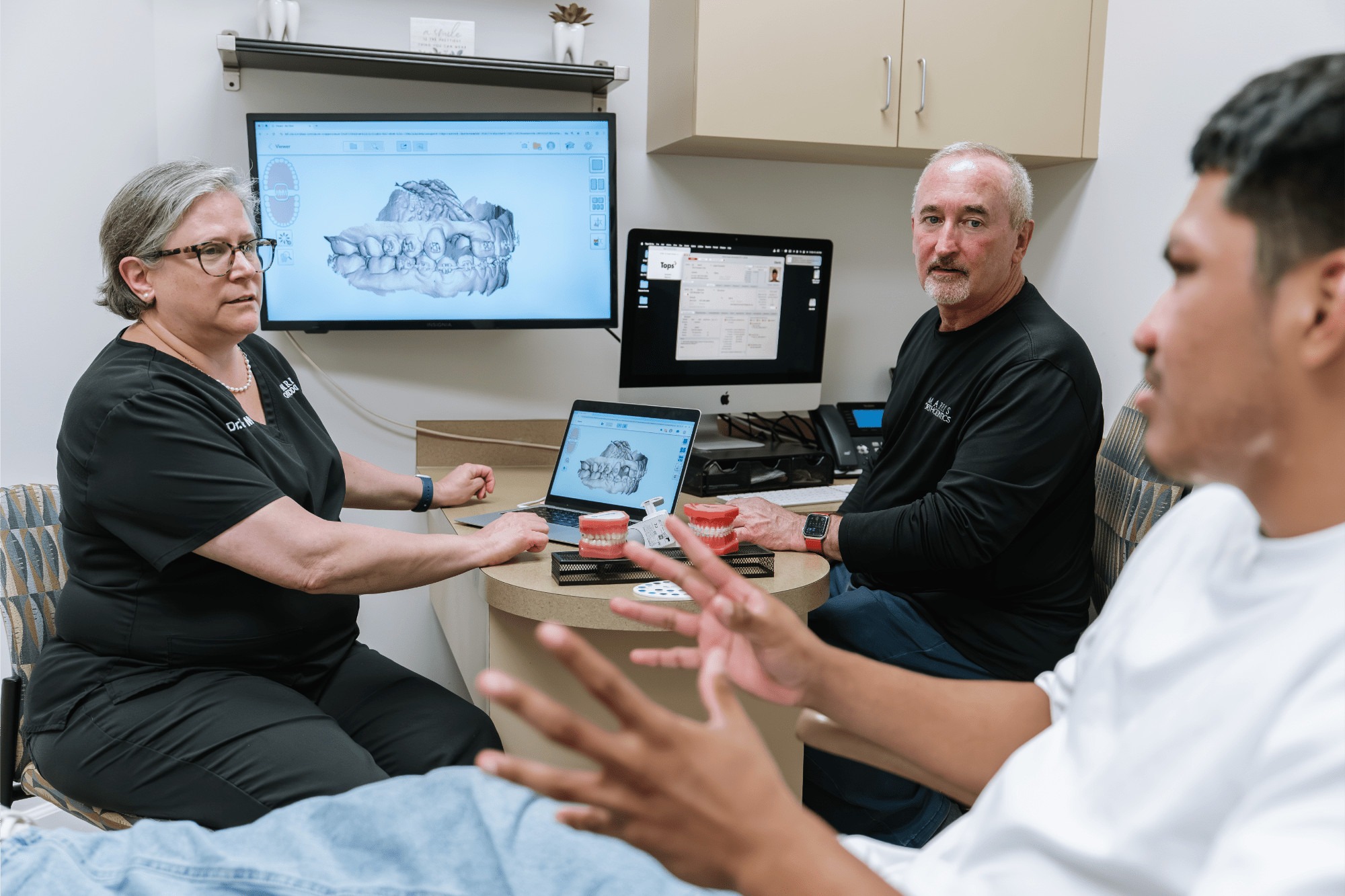 Orthodontic consultation at Marris Orthodontics, featuring Dr. Marris and staff discussing Invisalign treatment with a patient, digital scans displayed on screens, and orthodontic models on the table.