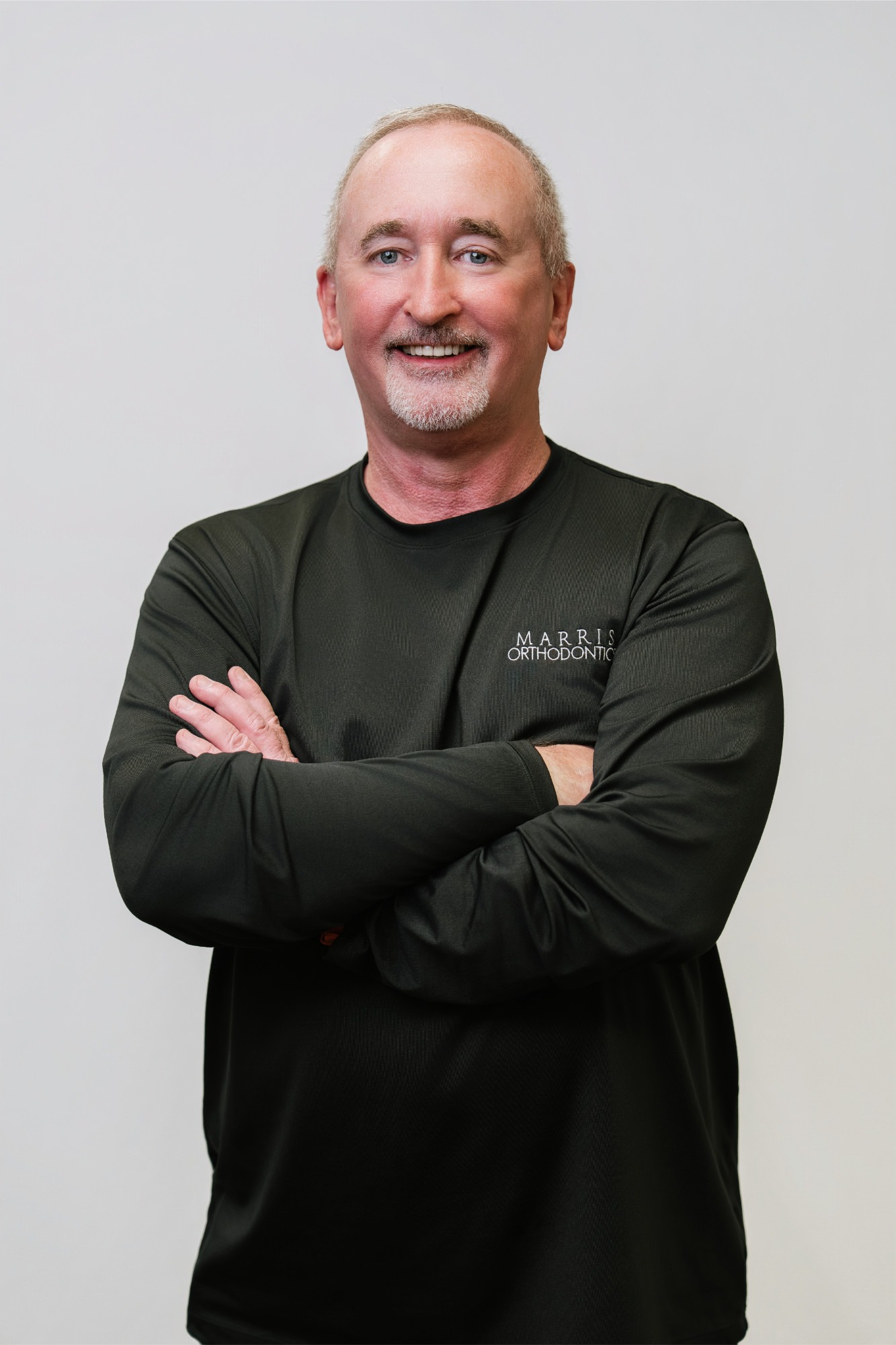 Man with crossed arms, smiling, wearing a black long-sleeve shirt with "Marris Orthodontics" logo, representing the orthodontic team at Marris Orthodontics, a leading provider of braces and Invisalign in Orlando.