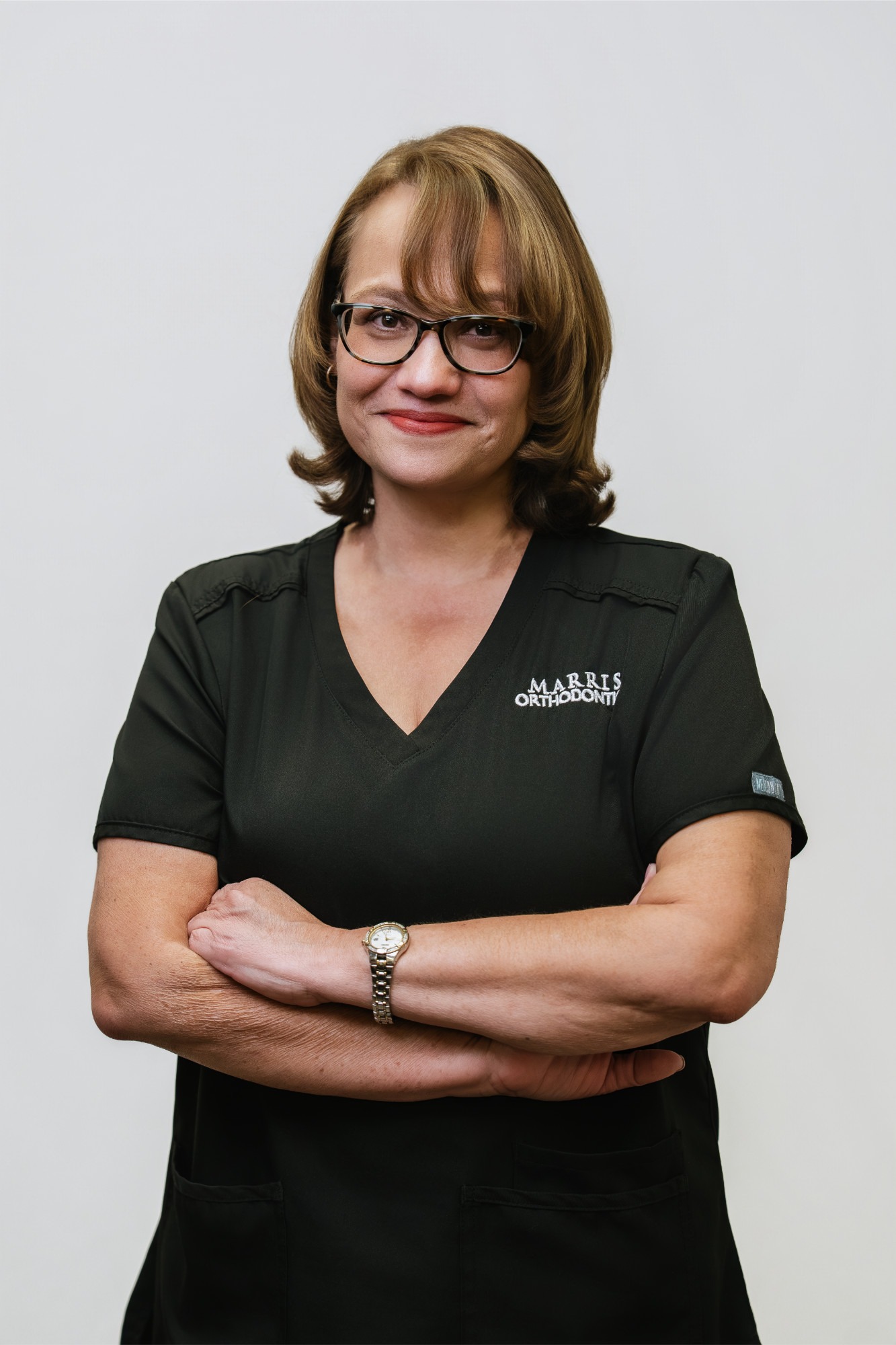 Nicki, Practice Administrator at Marris Orthodontics, wearing a black uniform with the brand logo, smiling and standing with arms crossed against a plain background.
