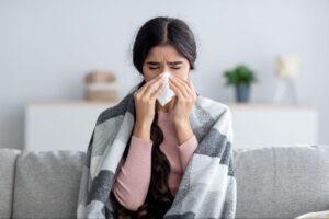 Young woman wrapped in a blanket, feeling unwell, holding a tissue to her nose, representing challenges of managing braces during illness, relevant to orthodontic care by Marris Orthodontics in Orlando.