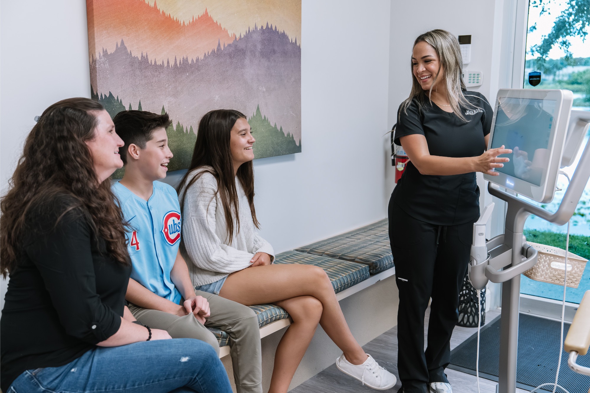 Marris Orthodontics team member showing a family their teen’s treatment plan on a digital touchscreen while a mom, a teen boy and a teen girl watch from a bench at our Orlando orthodontics office.