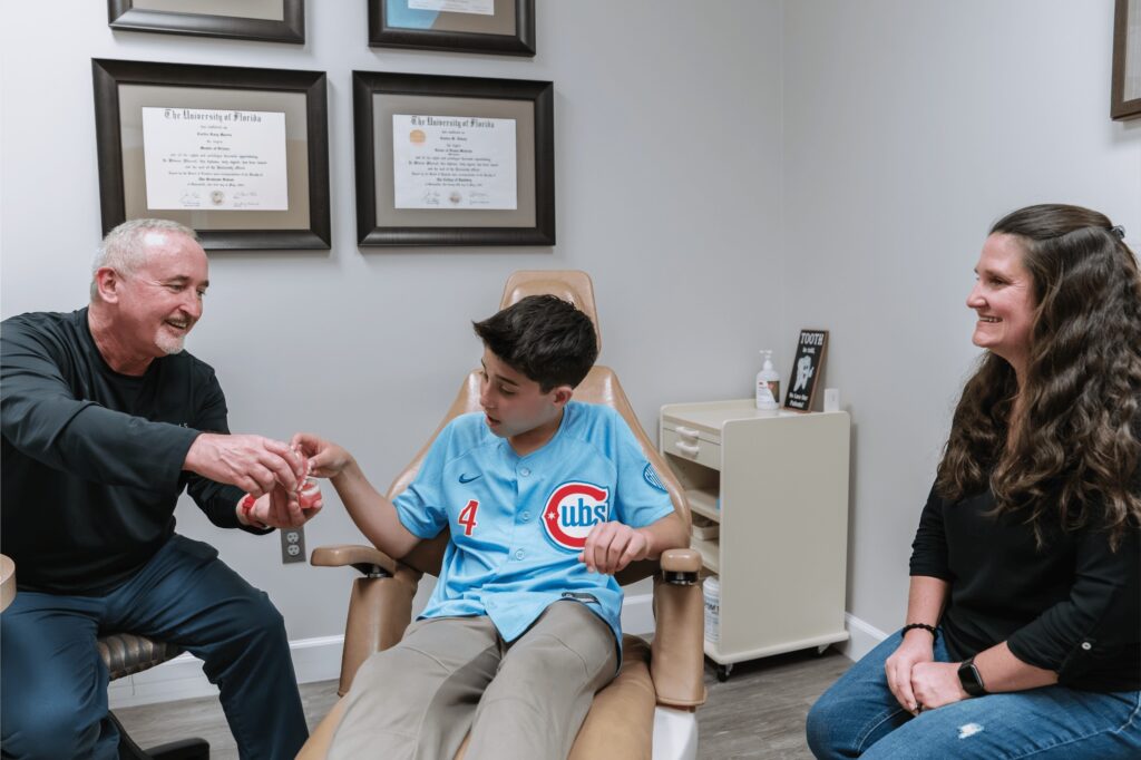 Orthodontic team member shows a teen boy a model of braces on teeth in a treatment chair at Marris Orthodontics in Orlando, FL, as his mom looks on