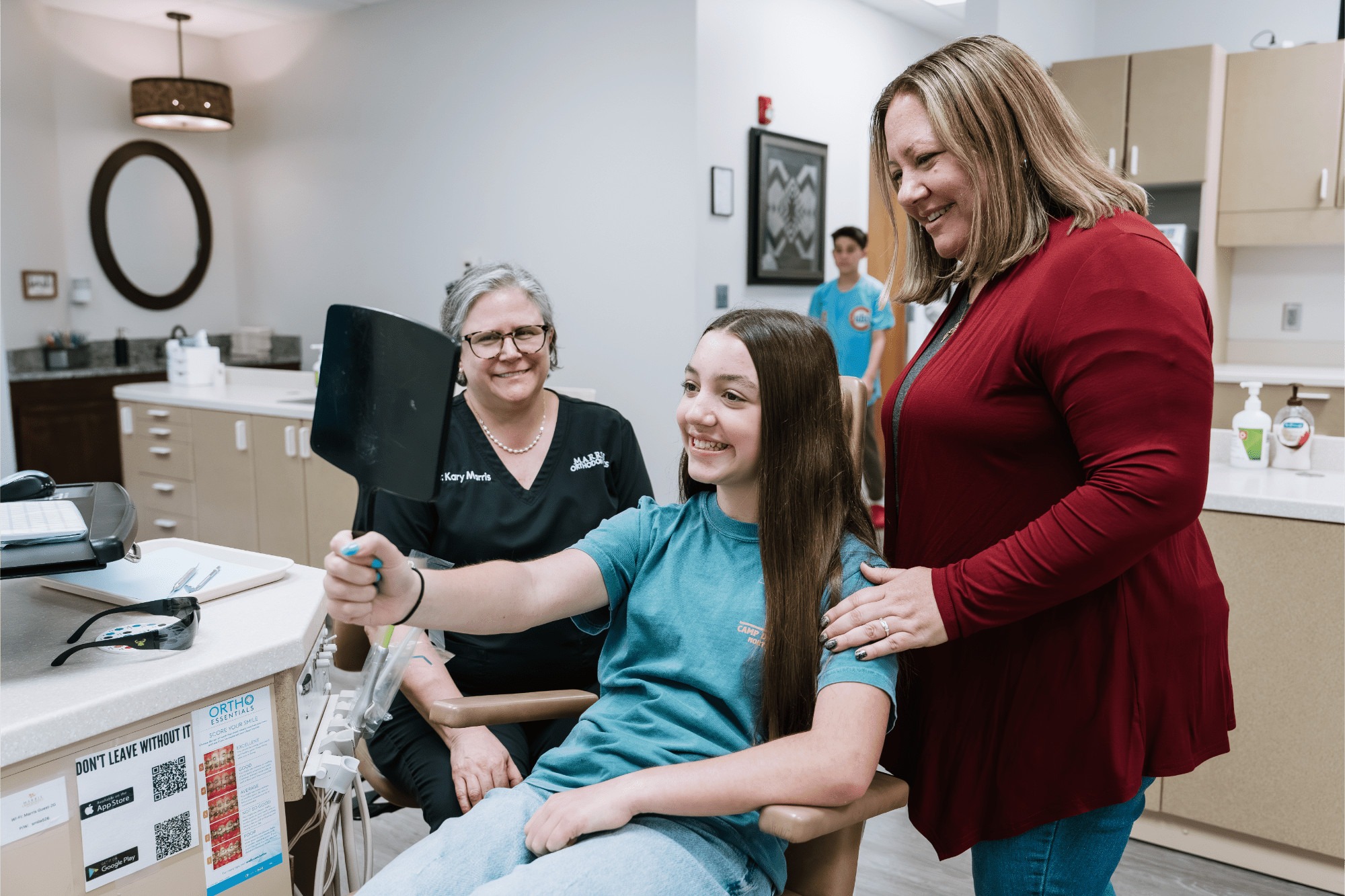 Dr. Kary Marris, Orlando orthodontist, smiling beside a teen girl in the treatment chair as she checks her new smile in a hand mirror while her mom rests a hand on her shoulder at Marris Orthodontics in Orlando, FL.