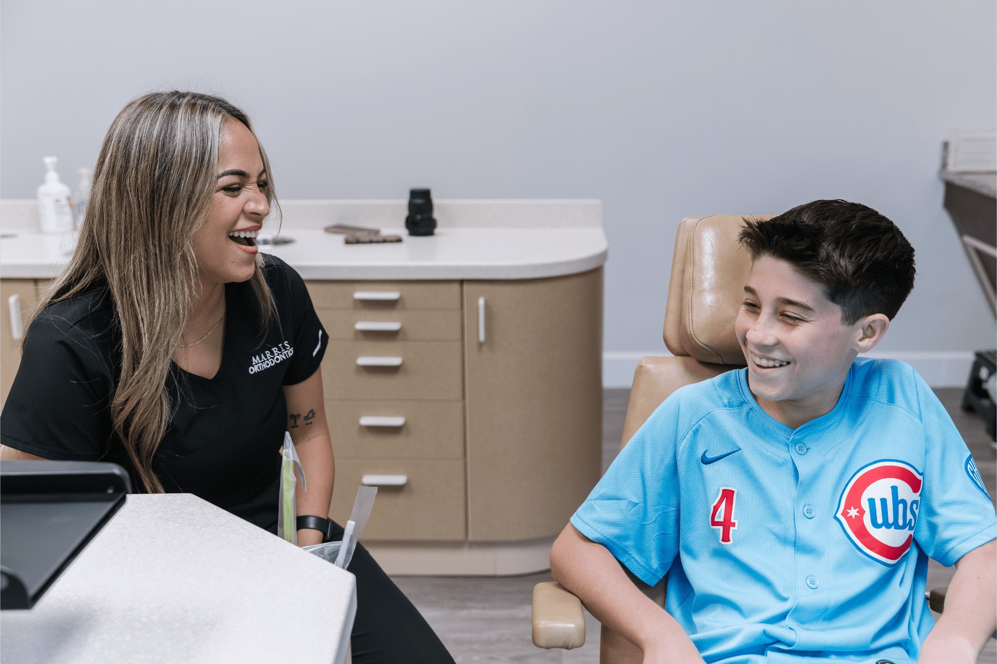 Smiling Marris Orthodontics team member laughing with a teen boy in a light blue baseball jersey as he sits in the treatment chair during his orthodontic visit at our Orlando orthodontics office.
